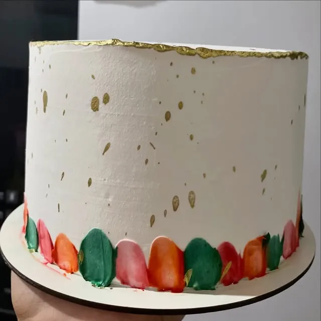 Bolo branco com borda dourada, respingos dourados e pinceladas coloridas na base